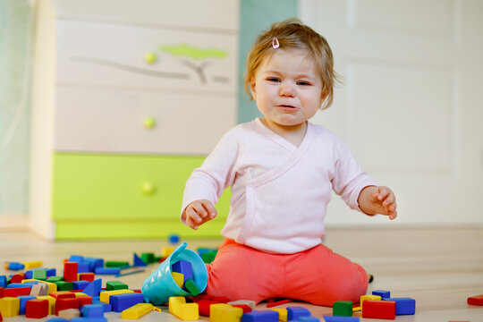 Upset Crying Baby Girl With Educational Toys. Sad Tired Or Hungry Alone Healthy Child Sitting Near Colorful Different Wooden Blocks At Home Or Nursery. Baby Missing Mother In Daycare
