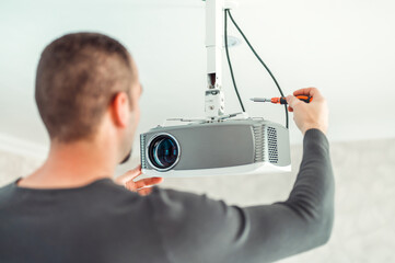 The man independently adjusts a multimedia video projector for home theater or presentations mounted on a ceiling bracket. - obrazy, fototapety, plakaty