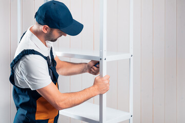 A uniformed worker is assembling a white metal shelving against a white clapboard wall.
