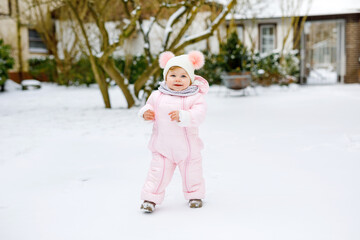 Happy little baby girl making first steps outdoors in winter through snow. Cute toddler learning walking. Child having fun on cold snowy day. Wearing warm baby pink clothes snowsuit and bobbles hat.