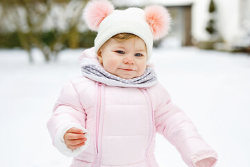 Happy little baby girl making first steps outdoors in winter through snow. Cute toddler learning walking. Child having fun on cold snowy day. Wearing warm baby pink clothes snowsuit and bobbles hat.