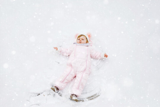 Cute little baby girl in colorful winter clothes making snow angel, laying down on snow. Active outdoors leisure with children in winter. Toddler in pink snowsuit.