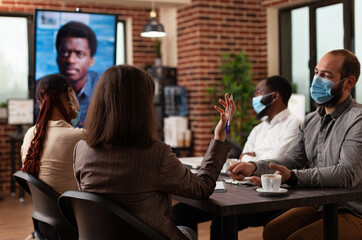 Multiethnic businesspeople with medical face mask to prevent infection with covid19 discussing company strategy with remote manager during online videocall meeting in startup office. Business call
