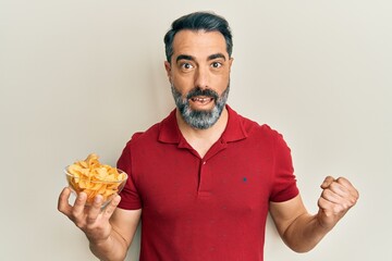 Middle age man with beard and grey hair holding potato chip screaming proud, celebrating victory and success very excited with raised arm