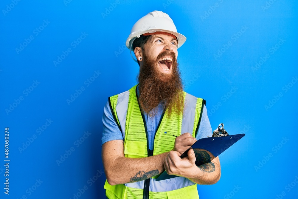 Poster redhead man with long beard wearing safety helmet holding clipboard angry and mad screaming frustrat