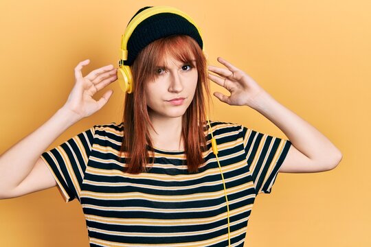 Redhead young woman listening to music using headphones clueless and confused expression. doubt concept.