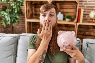 Redhead man holding piggy bank covering mouth with hand, shocked and afraid for mistake. surprised...
