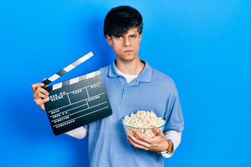 Handsome hipster young man eating popcorn holding cinema clapboard skeptic and nervous, frowning...