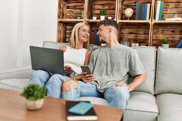 Young caucasian couple smiling happy using laptop and smartphone sitting on the sofa at home.