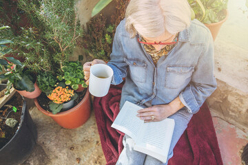 Elderly woman reads book surrounded by plants with cup in her hand