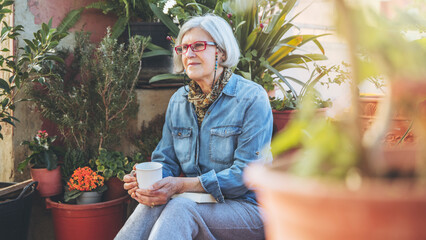 Thoughtful older woman, sitting between plants, holding cup