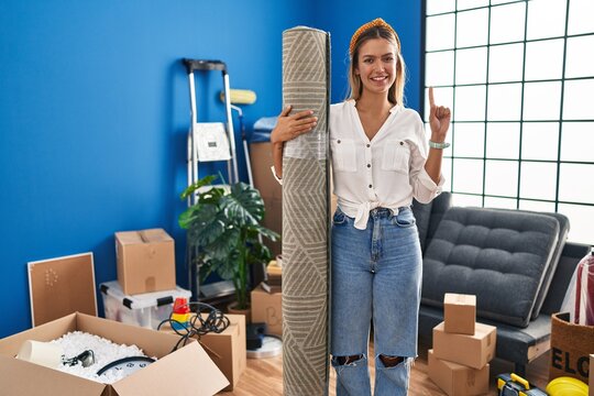 Young blonde woman moving to a new home holding rug smiling with an idea or question pointing finger with happy face, number one