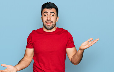 Hispanic man with beard wearing casual red t shirt clueless and confused expression with arms and...