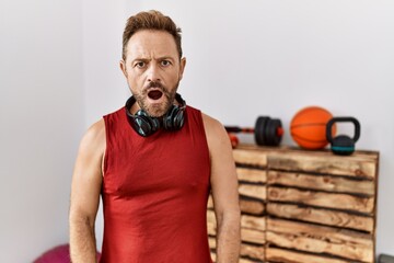 Middle age man wearing sportswear and headphones at the gym scared and amazed with open mouth for...