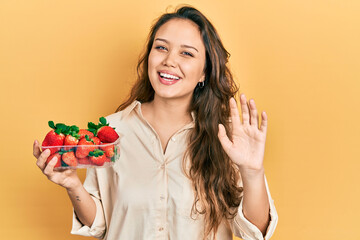 Young hispanic girl holding strawberries waiving saying hello happy and smiling, friendly welcome...