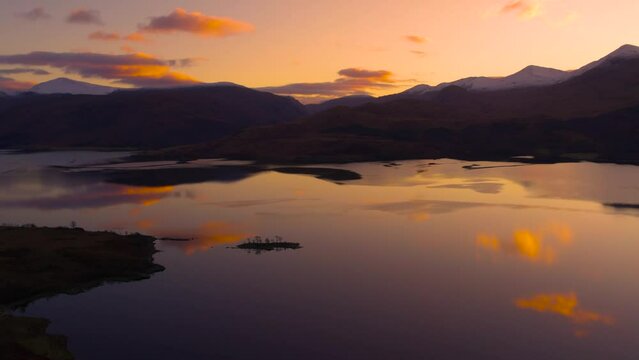 Aerial drone footage panning slowly to reveal Glen Etive and Loch Etive in Scotland during an orange and pink sunrise of pastel colours, with snow-capped mountains, trees and still reflective water.