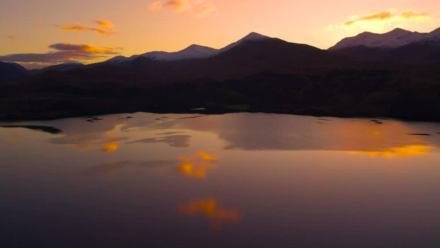 Slow reverse pan aerial drone footage revealing Glen Etive and Loch Etive in Scotland during an orange and pink sunrise of pastel colours, with snow-capped mountains, trees and still reflective water.