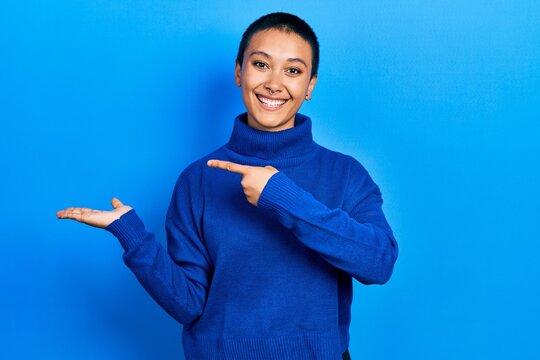 Beautiful hispanic woman with short hair wearing turtleneck sweater amazed and smiling to the camera while presenting with hand and pointing with finger.