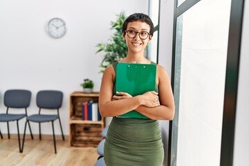 Young hispanic woman smiling confident holding clipboard standing at waiting room