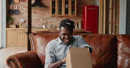 Emotional excited young Black guy unpack small box parcel feel surprised overjoyed with gift send by friend. Amazed male customer get awaited lottery prize giveaway present by courier delivery service