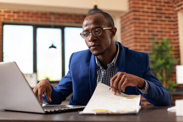 Fototapeta premium Employee working with laptop and papers on clipboard to plan business project. Man using computer and analyzing documents for financial strategy at startup office. Entrepreneur at work