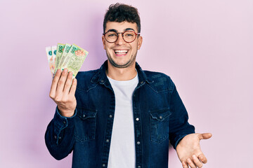 Young hispanic man holding argentine pesos banknotes celebrating achievement with happy smile and...