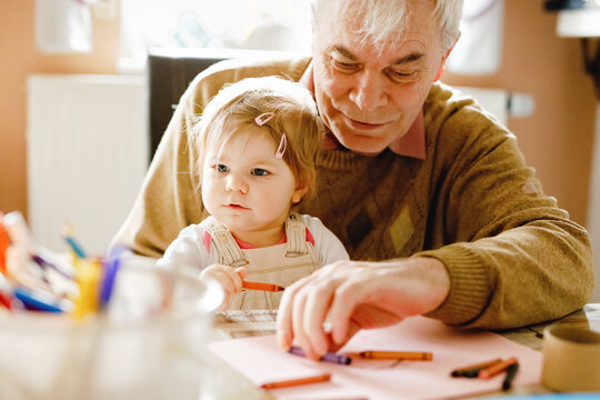 Cute little baby toddler girl and handsome senior grandfather painting with colorful pencils at home. Grandchild and man having fun together. Family and generation in love