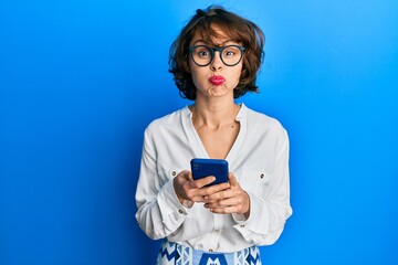 Fototapeta premium Young brunette woman using smartphone puffing cheeks with funny face. mouth inflated with air, catching air.