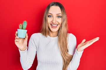 Young blonde girl holding small cactus pot celebrating victory with happy smile and winner expression with raised hands