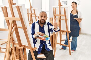 Senior artist man at art studio approving doing positive gesture with hand, thumbs up smiling and happy for success. winner gesture.