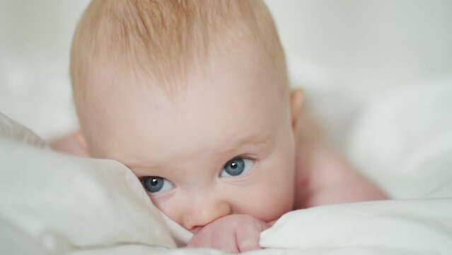 Cute baby lying on bed. Adorable infant with gray eyes sucking blanket and yawning while resting on soft bed at home