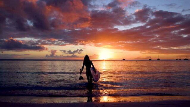 A woman with a stand up paddle board (SUP) walks into the calm sea during a colorful sunset in the Caribbean