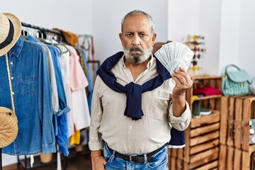 Handsome senior man at retail shop holding dollars banknotes thinking attitude and sober expression looking self confident