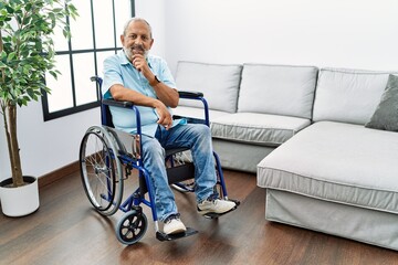 Handsome senior man sitting on wheelchair at the living room looking confident at the camera smiling with crossed arms and hand raised on chin. thinking positive.