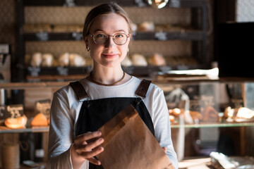 Young smiling woman (salesperson in bakery) holding craft paper packing bag for taking away food in...