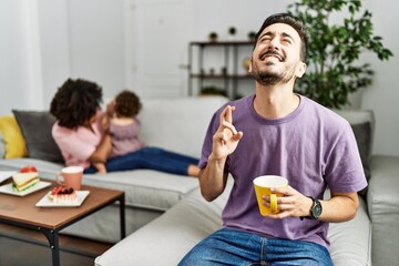 Hispanic father of interracial family drinking a cup coffee gesturing finger crossed smiling with hope and eyes closed. luck and superstitious concept.