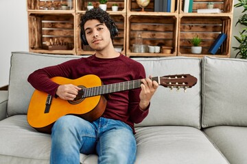 Young hispanic man playing classical guitar sitting on the sofa at home.
