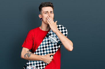 Young caucasian boy with ears dilation holding skate covering mouth with hand, shocked and afraid...