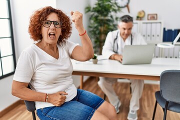 Senior woman sitting at doctor appointment angry and mad raising fist frustrated and furious while...