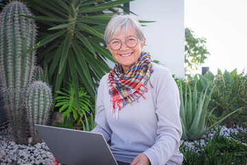 Happy mature senior woman smiling looking at camera while using laptop computer outside home sitting in the garden. Active elderly carefree people enjoying tech and social. Travel or retirement