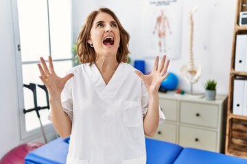 Middle age physiotherapist woman working at pain recovery clinic crazy and mad shouting and yelling with aggressive expression and arms raised. frustration concept.