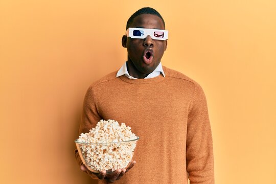 Young african american man wearing d glasses holding popcorns scared and amazed with open mouth for surprise, disbelief face