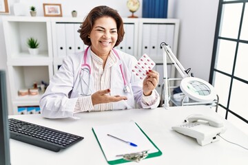 Middle age hispanic woman wearing doctor uniform holding pills at clinic