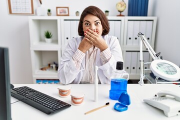 Middle age hispanic dentist woman working at medical clinic shocked covering mouth with hands for mistake. secret concept.