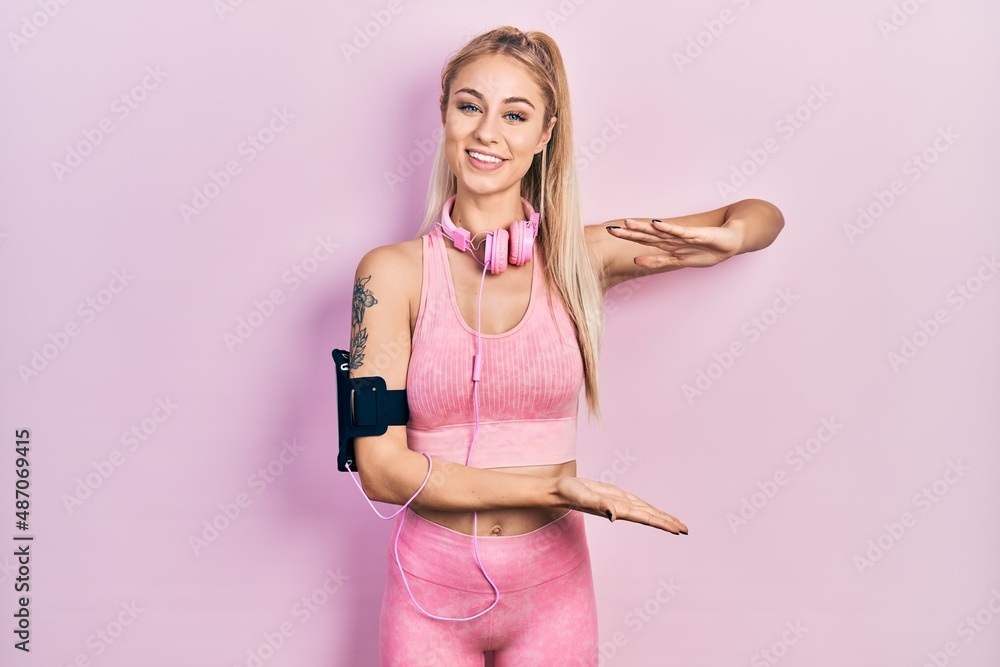 Poster young beautiful caucasian woman wearing gym clothes and using headphones gesturing with hands showin