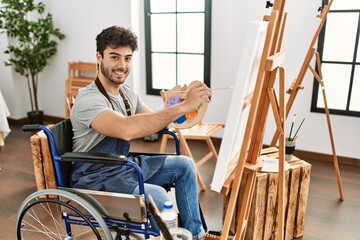 Young hispanic artist disabled man smiling happy painting at art studio.