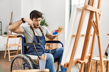 Young hispanic man sitting on wheelchair painting at art studio pointing down with fingers showing...