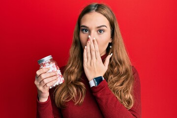Young blonde woman holding jar of pills covering mouth with hand, shocked and afraid for mistake....