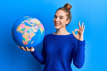 Young blonde woman holding world ball doing ok sign with fingers, smiling friendly gesturing excellent symbol
