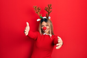 Beautiful hispanic woman wearing deer christmas hat and red nose looking at the camera smiling with open arms for hug. cheerful expression embracing happiness.